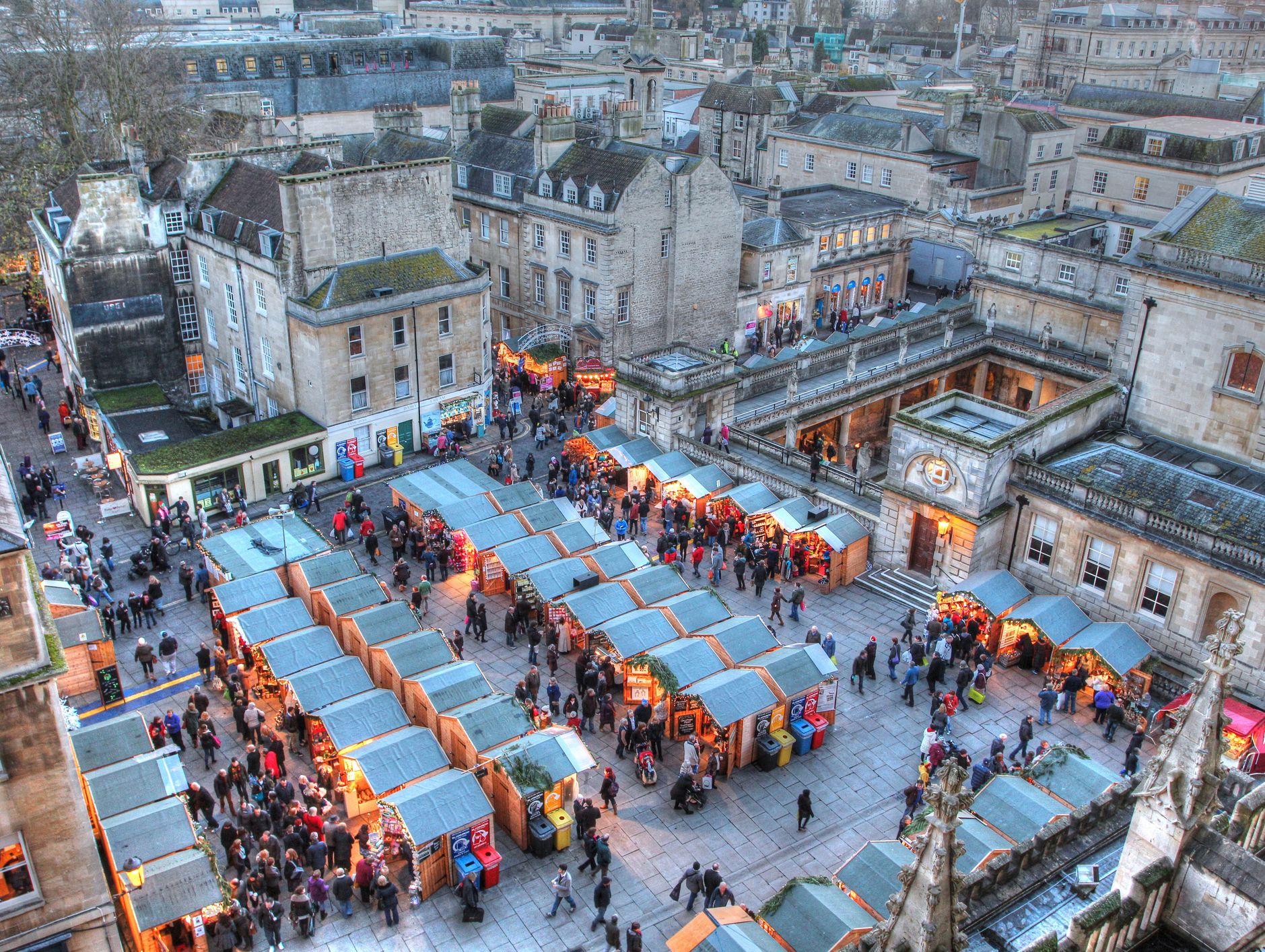 A bustling Christmas market in Wiltshire, featuring rows of warmly lit wooden stalls, surrounded by historic buildings, and lively crowds enjoying festive shopping and activities.