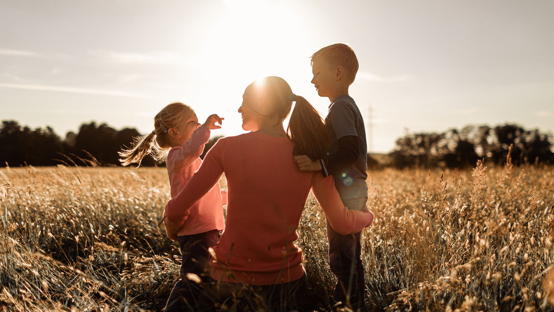 A mother kneels in a sunlit field, embracing her two young children as they laugh and play together, with the warm glow of the sunset behind them.