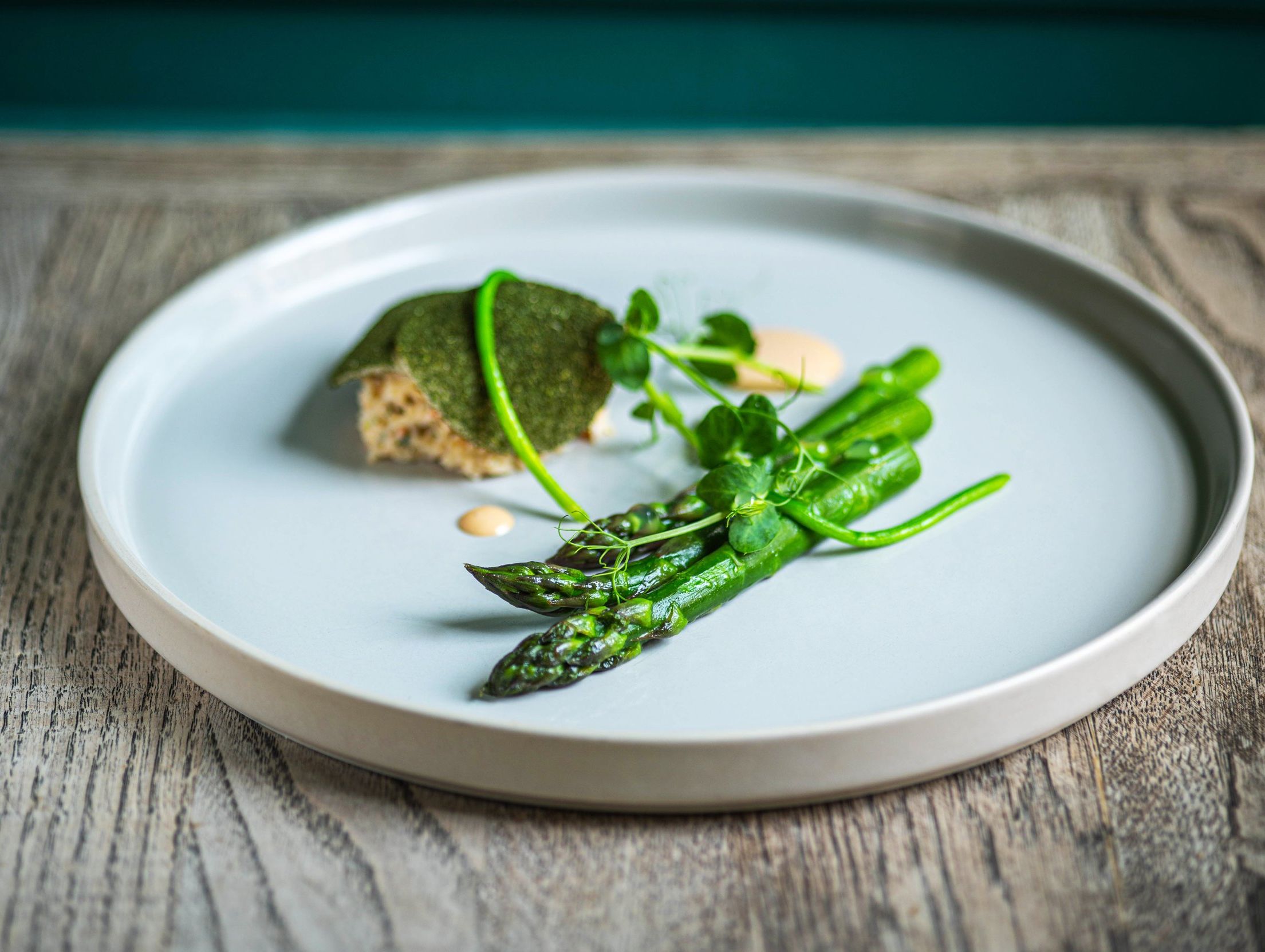A minimalist gourmet dish featuring asparagus, pea shoots, and a delicate portion of fish topped with a green crust, served on a light ceramic plate on a wooden table.