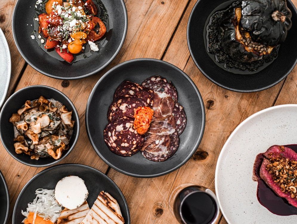 An assortment of gourmet dishes served in dark plates, including cured meats, roasted vegetables, grilled bread, and vibrant salads, arranged on a rustic wooden table.