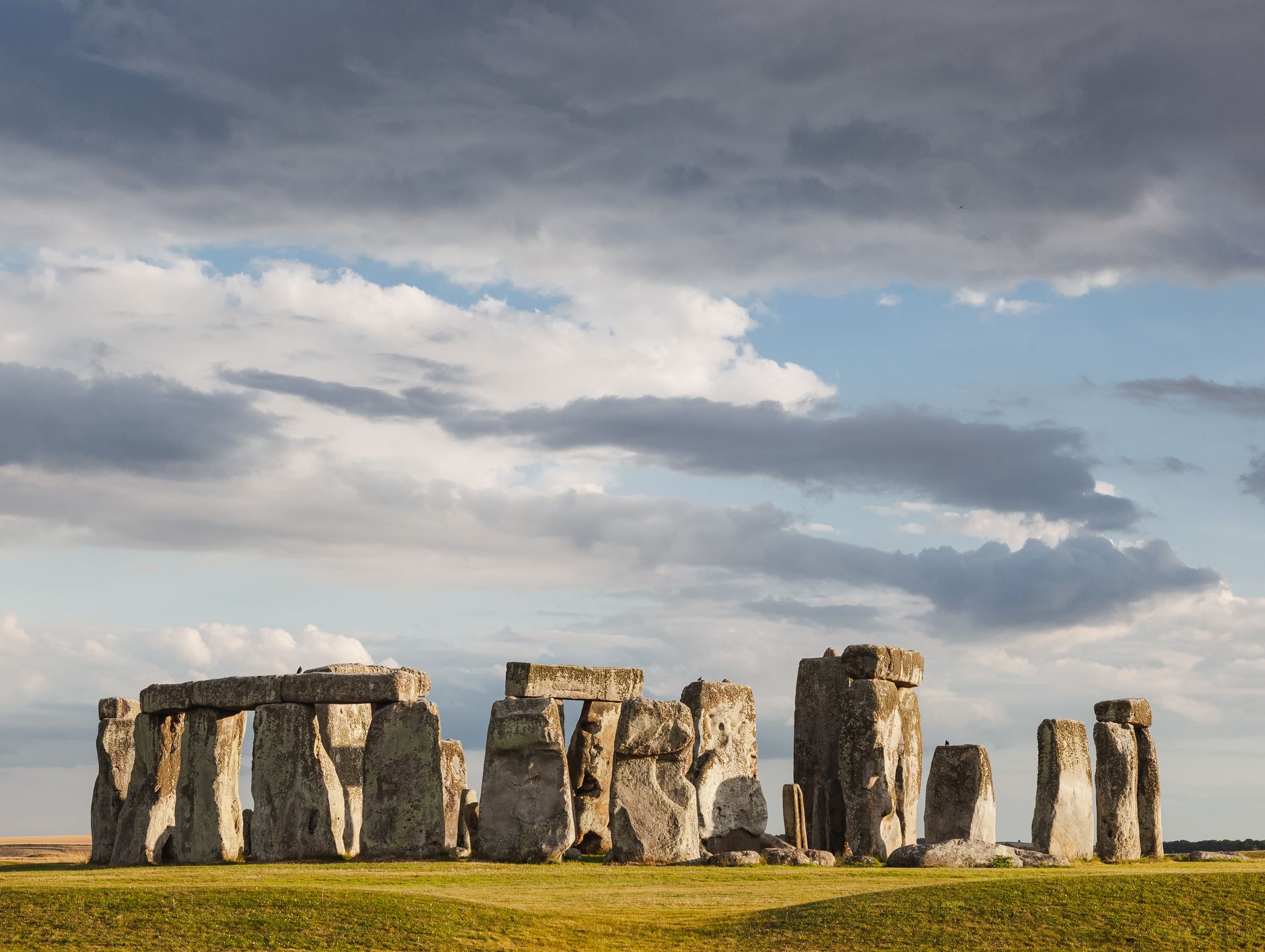 The iconic Stonehenge stone circle stands under a dramatic, partly cloudy sky, with soft light casting shadows on the grassy field surrounding the ancient monument.