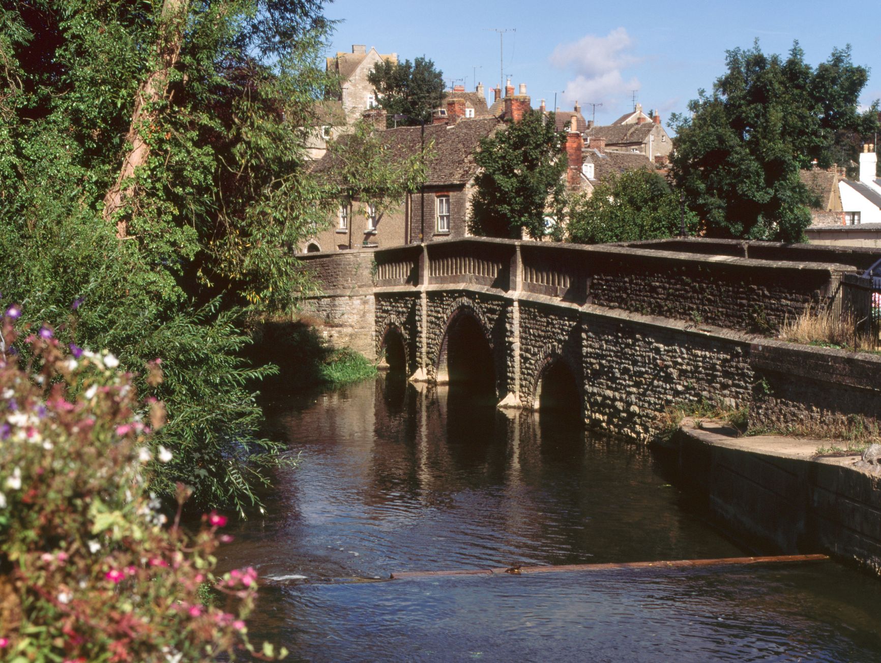 Malmesbury bridge with multiple arches spans a calm river, surrounded by greenery and flowers in the foreground, with a village of brick houses visible in the background.