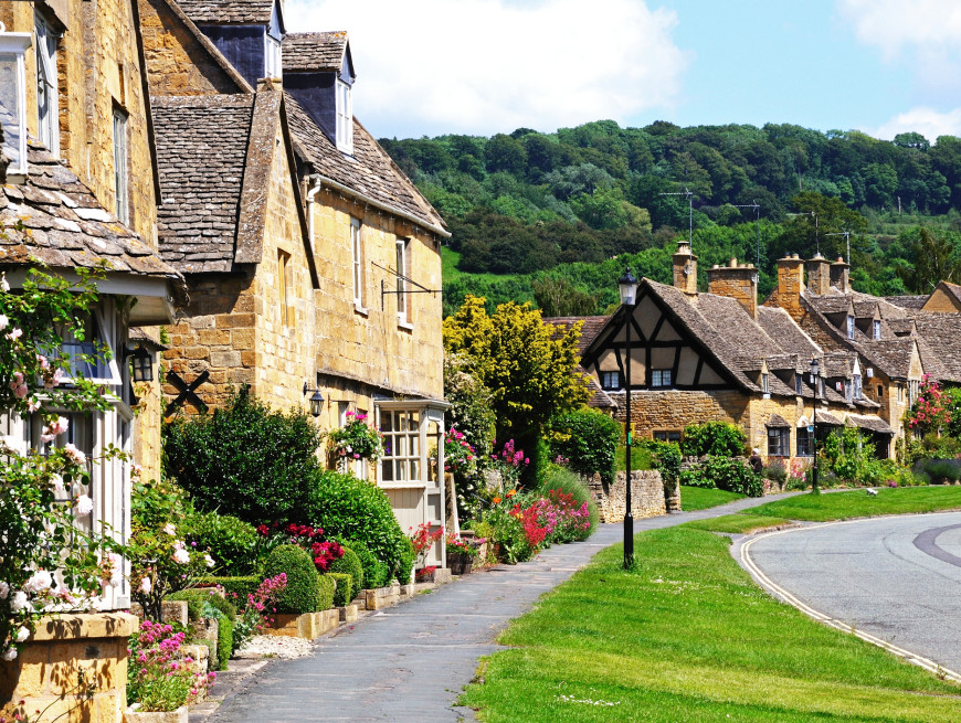 A charming village street lined with stone cottages, blooming flower gardens, and greenery, with a backdrop of lush, wooded hills under a bright blue sky with fluffy clouds.