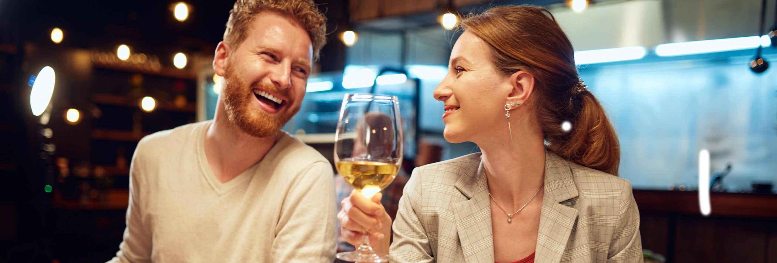 A smiling couple enjoying dinner at a restaurant, with the woman raising a glass of white wine and plates of food, including fries and appetizers, on a wooden table.
