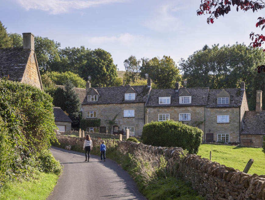 A quiet village lane bordered by stone cottages and lush greenery, where a woman and child walk along the road with a small dog under a bright, clear sky in a serene rural setting.