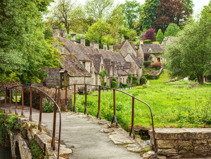 A charming village scene with stone cottages featuring sloped roofs, a narrow bridge crossing a small stream, and lush greenery surrounding the idyllic setting under a partly cloudy sky.