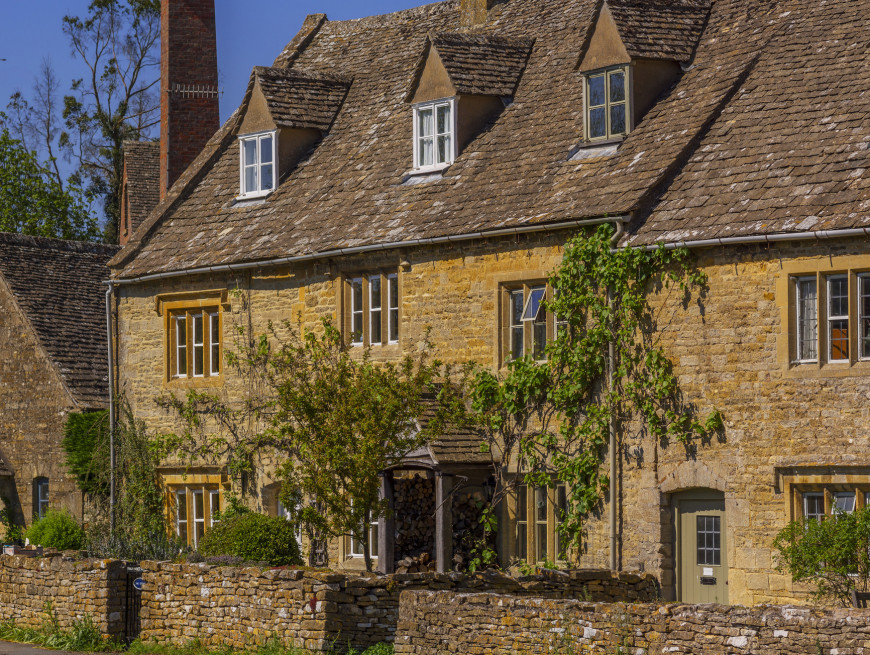 A traditional stone cottage with a sloped roof and multiple chimneys, adorned with climbing plants and surrounded by a low stone wall, set under a clear blue sky.