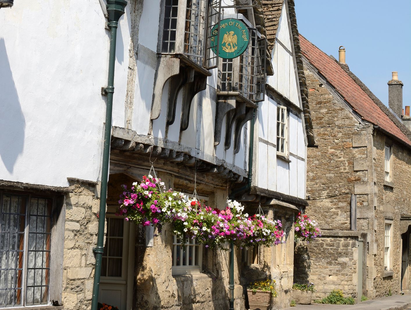 A historic building with whitewashed walls, timber framing, and a thatched roof stands on a quiet street. Hanging flower baskets and a pub sign add color to the scene.