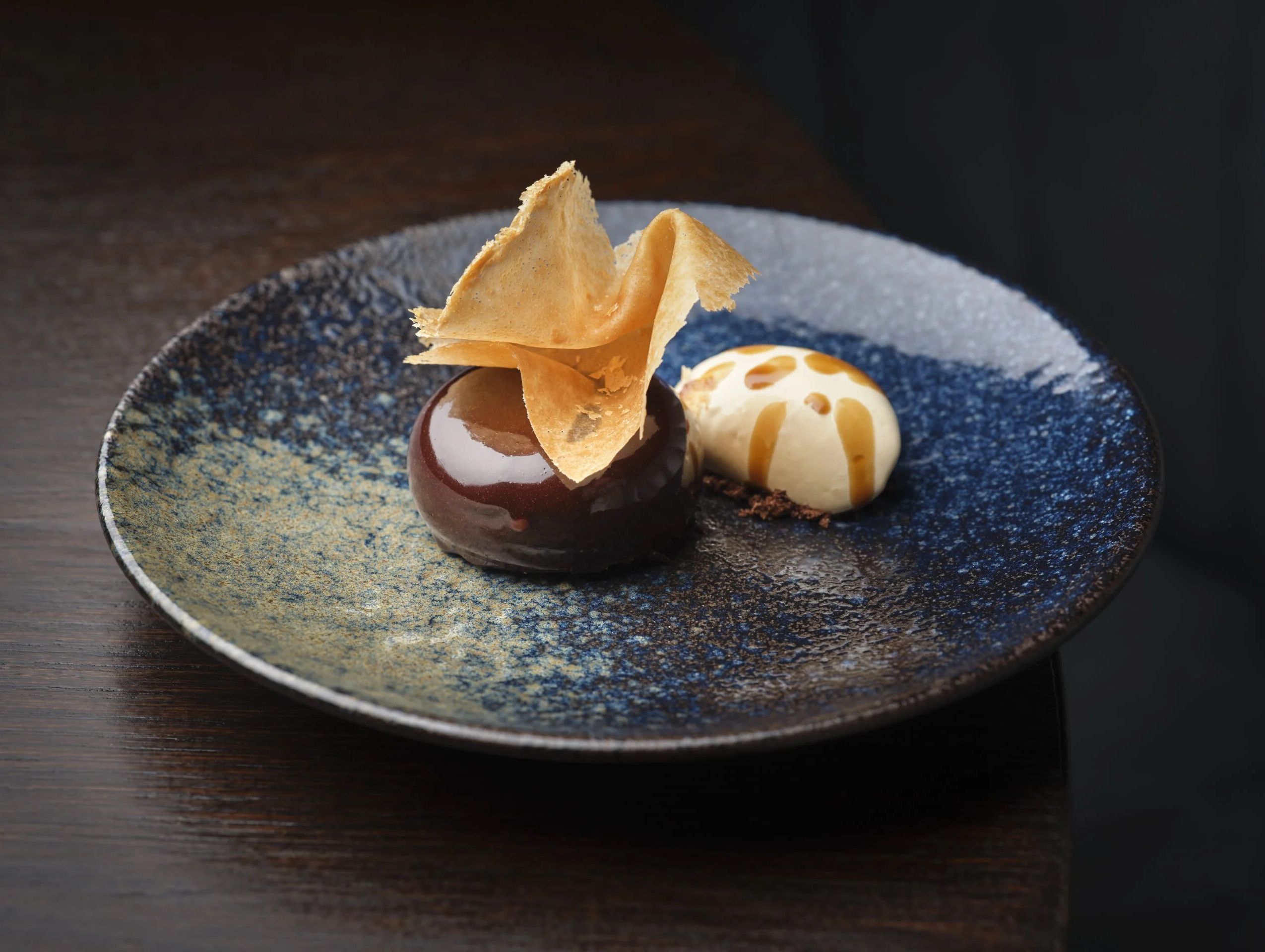 A beautifully plated dessert featuring a glossy chocolate sphere topped with a delicate pastry shard, served alongside a dollop of cream with caramel drizzle on a textured blue plate.