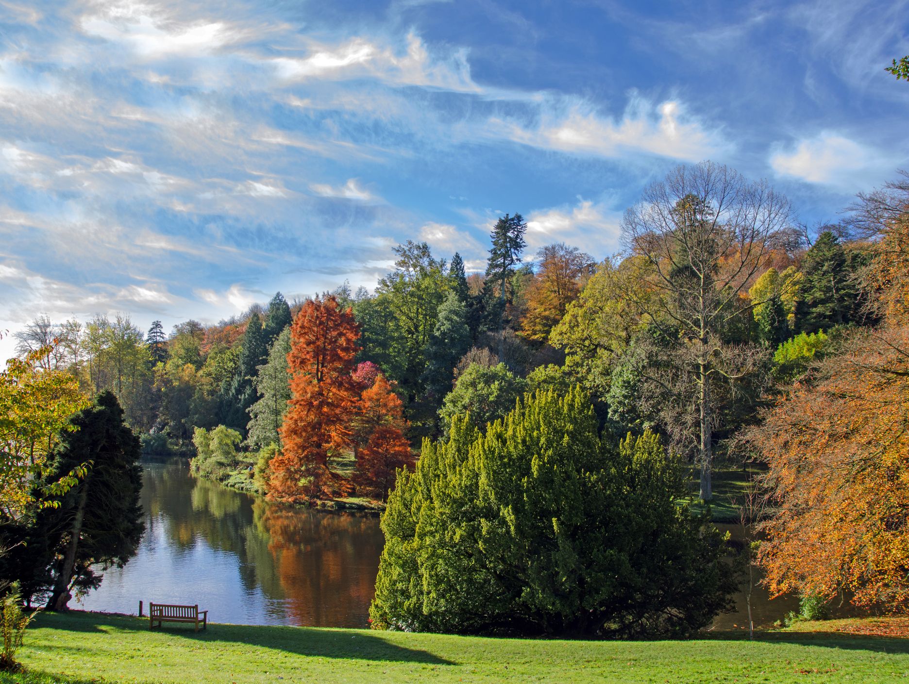 A serene landscape with autumn trees in vibrant colors reflecting on a calm lake, set under a blue sky with wispy clouds. A wooden bench overlooks the peaceful scene.