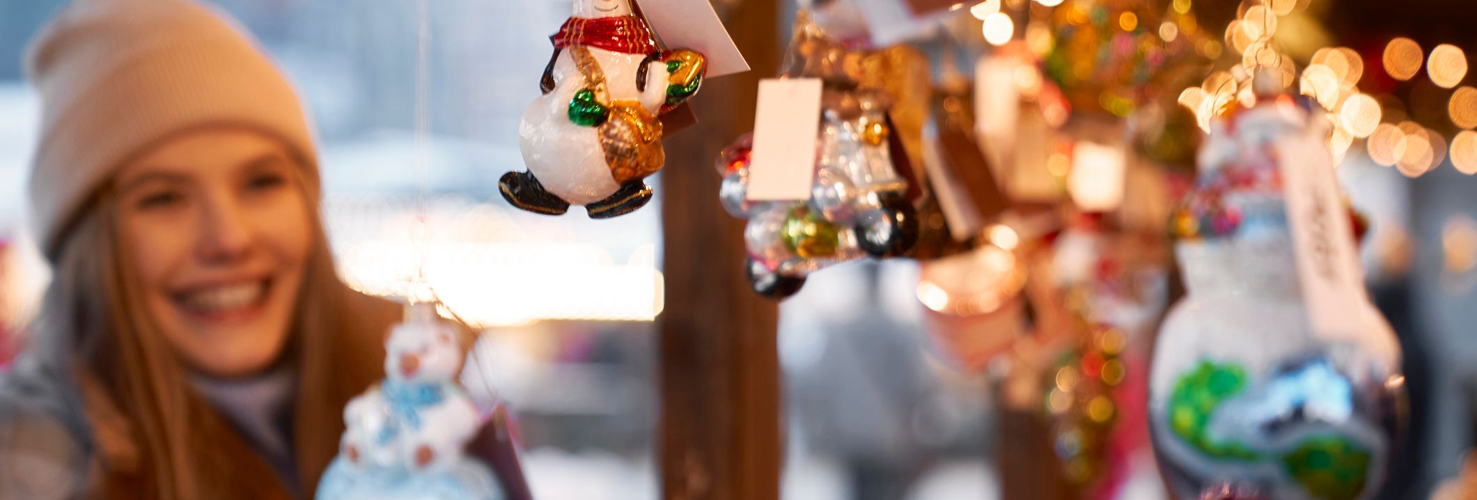 A smiling woman admires festive glass ornaments, including snowmen and baubles, at a twinkling Christmas market, capturing the joy of Christmas in Wiltshire.