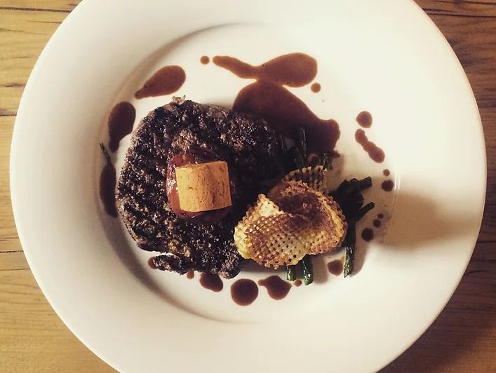 A top-down view of a gourmet dish featuring a seared steak topped with butter, crispy potato garnish, green beans, and drizzled brown sauce, served on a white plate over a wooden table.