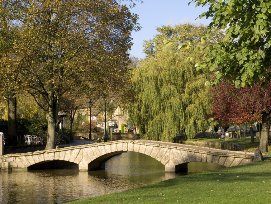 A charming stone footbridge spans a calm river in a park-like setting, surrounded by lush green grass and trees with autumn foliage, under a clear blue sky.