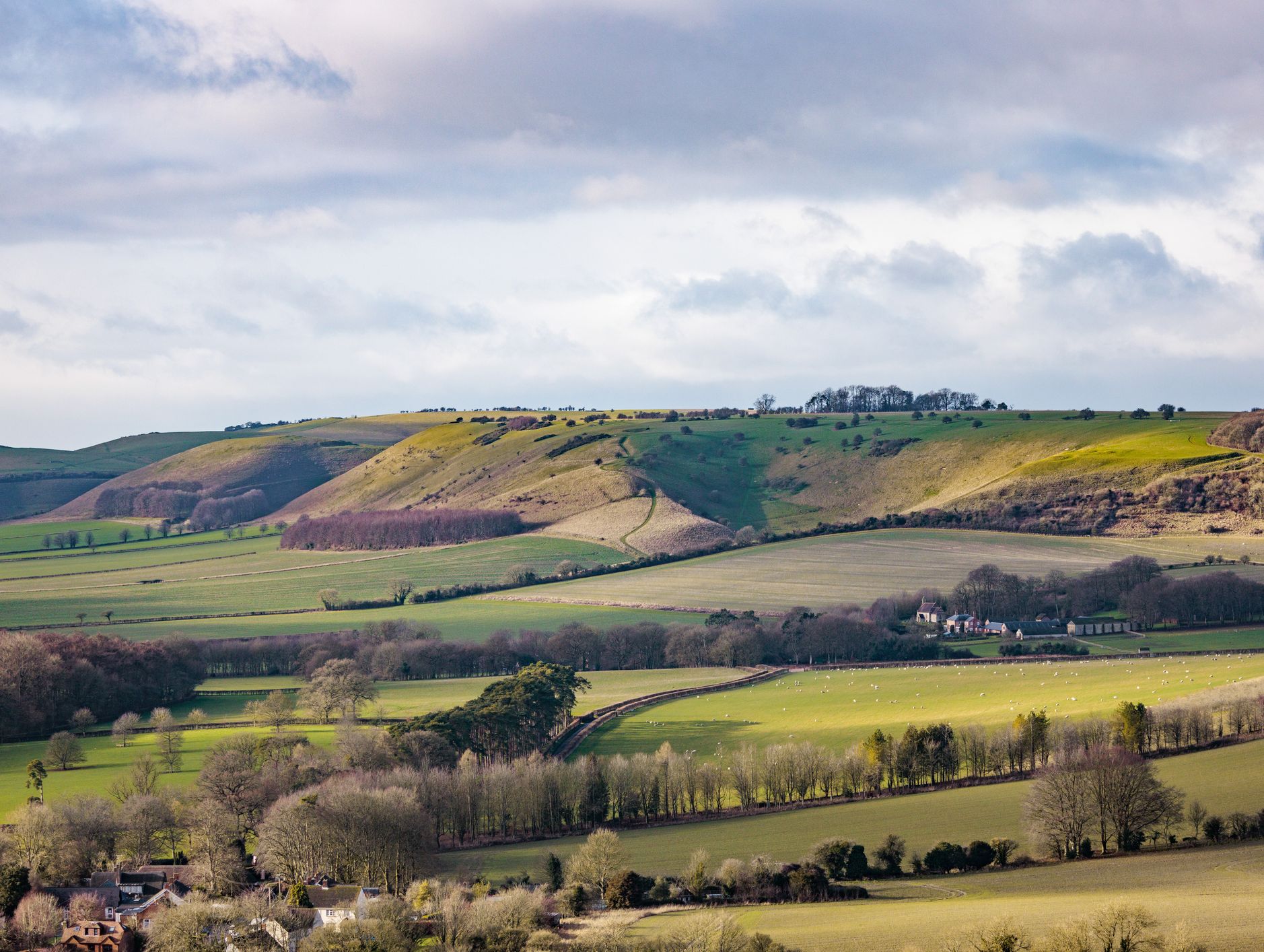 A scenic view of rolling green hills and farmland, with scattered trees and patches of sunlight breaking through clouds. A small village nestles at the base of the hills.
